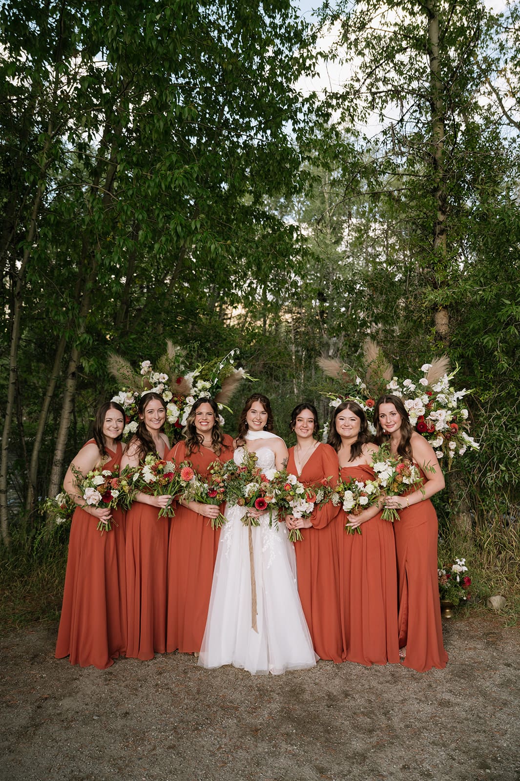 Five women stand together outdoors, one in a white wedding dress and four in matching rust-colored dresses, all holding bouquets of flowers and smiling.