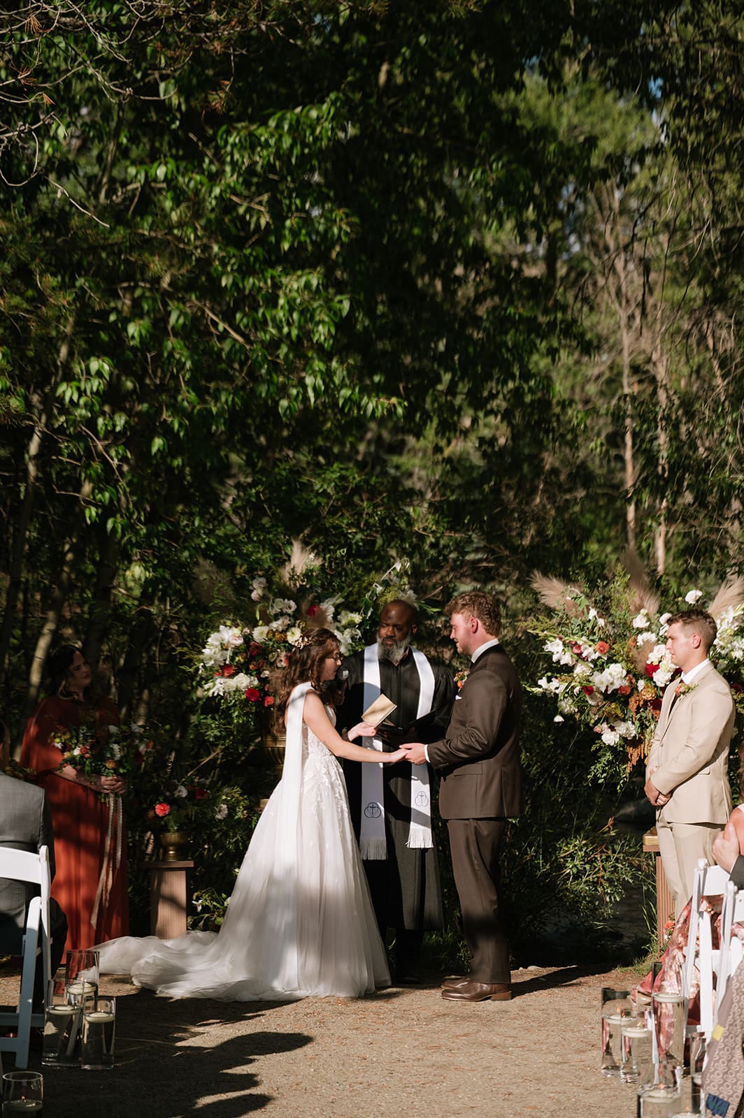 A bride and groom stand together during an outdoor Colorado wedding ceremony as the officiant speaks; groomsmen in beige suits stand in the background