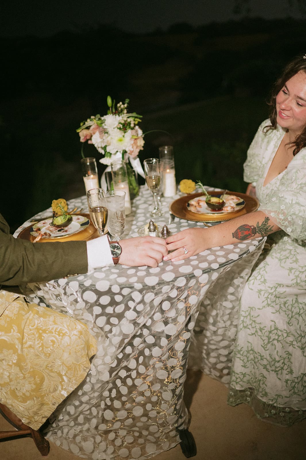 A couple sits at a small, elegantly set table outdoors, holding hands across the table, with plates of food and a floral centerpiece in view.