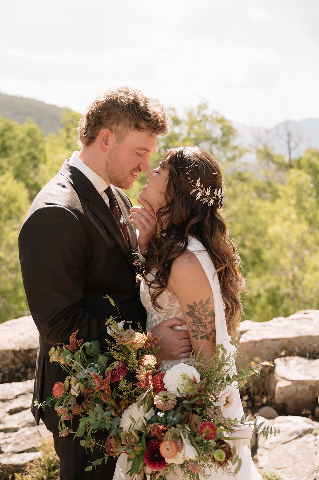 A bride and groom in wedding attire stand embracing on a rocky landscape with mountains and trees in the background under a partly cloudy sky for a Colorado wedding