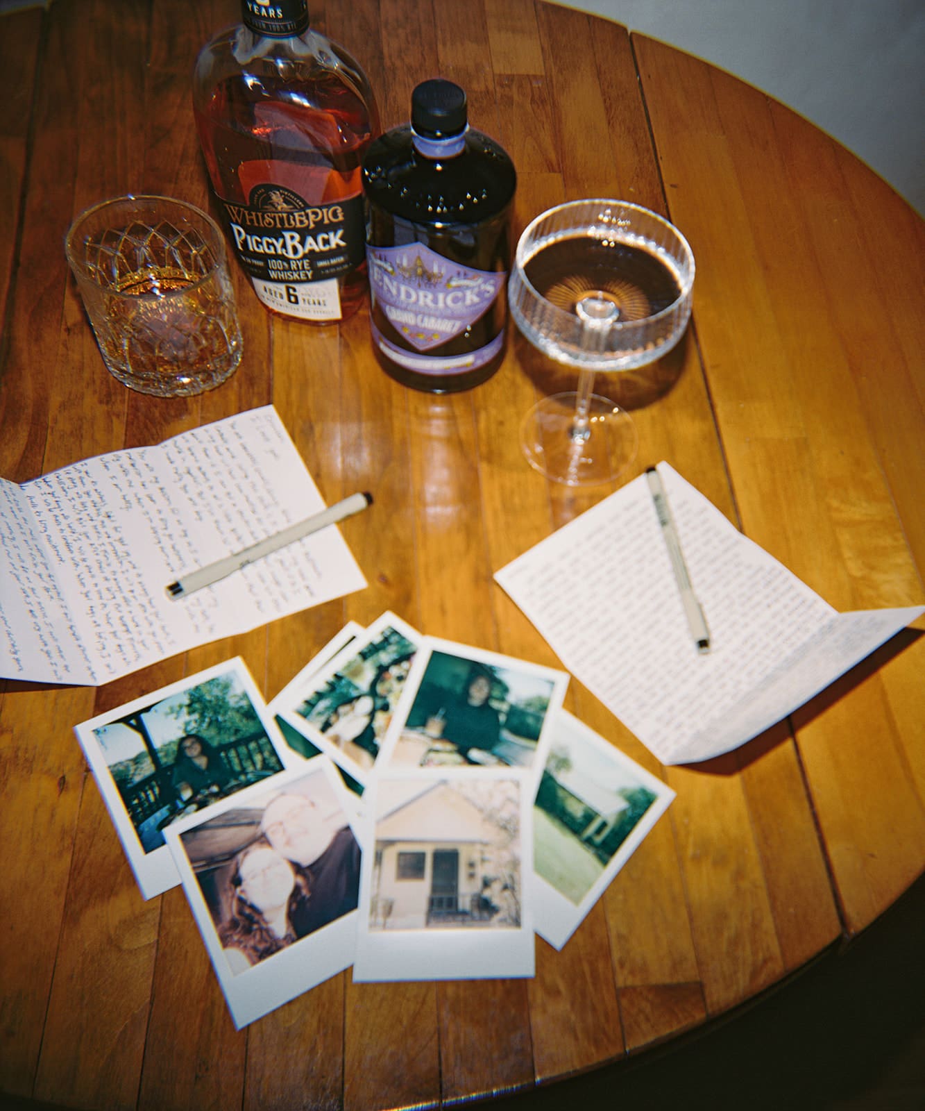 A wooden table with two handwritten notes, pens, a whiskey bottle, a gin bottle, a glass of whiskey, a cocktail glass, and several instant photos spread out.