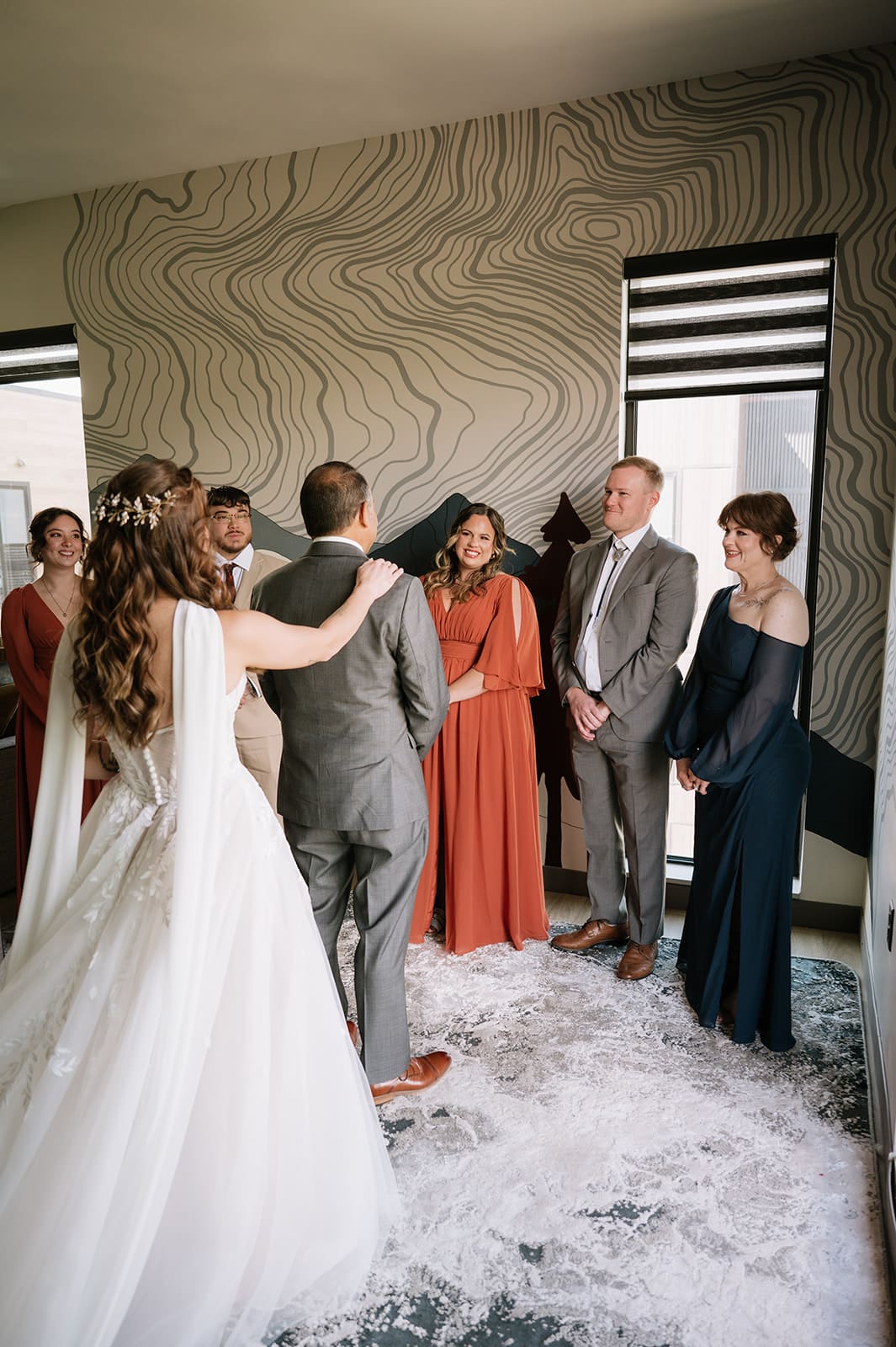A bride and groom stand together facing a group of people indoors, with floral petals on the floor and modern wall decor in the background.
