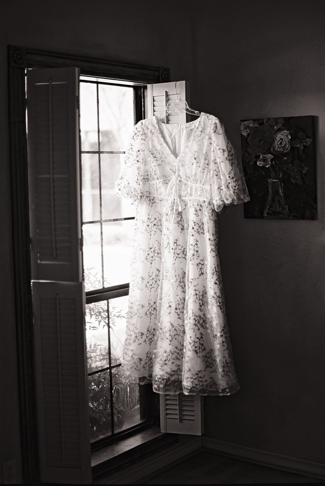 A white lace dress hangs on a hanger in front of a window with wooden shutters, letting in natural light.