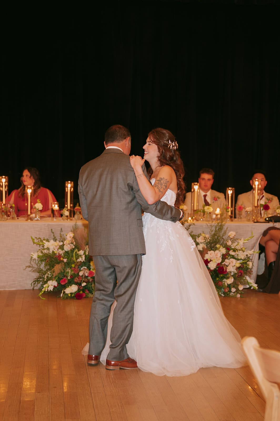 A bride and groom dance in the center of a large hall with guests seated at tables, string lights overhead, and a stage in the background.