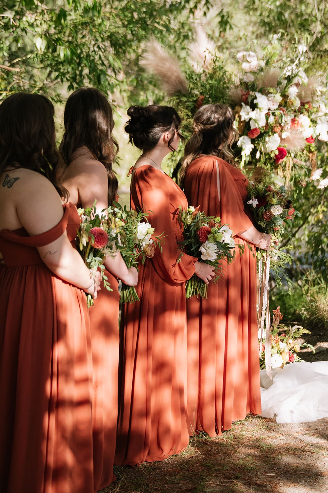 Four bridesmaids in matching burnt orange dresses stand outdoors holding bouquets at a wedding ceremony, with greenery and floral arrangements in the background.