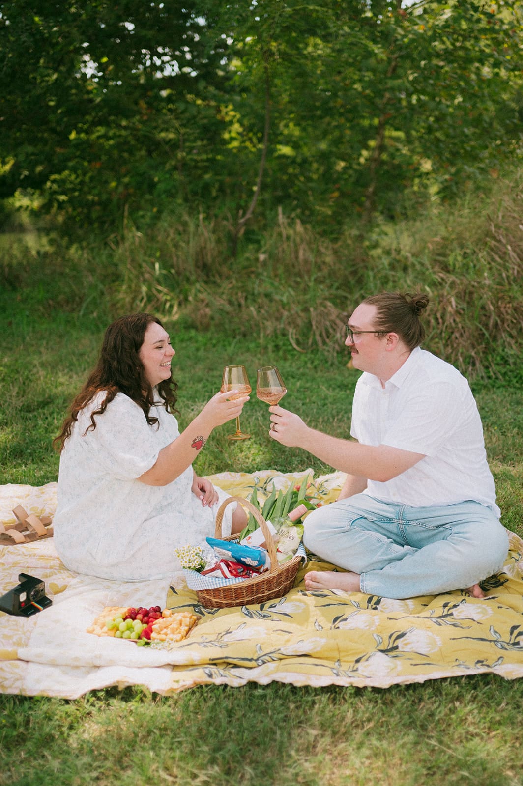 Two people sit on a blanket having a picnic outdoors, surrounded by greenery, with food and flowers arranged between them for a Texas Hill Country Elopement