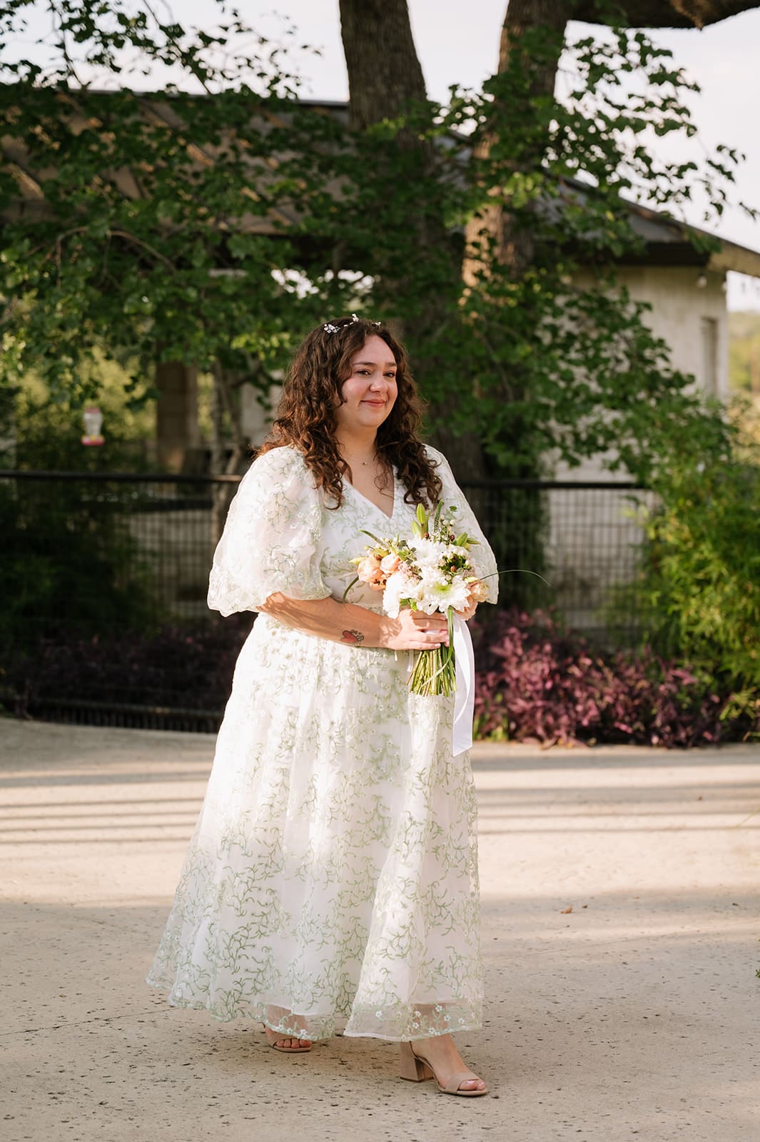 A woman in a white lace dress holding a bouquet walks outdoors on a sunny day, with greenery and a building in the background for a texas hill country elopement