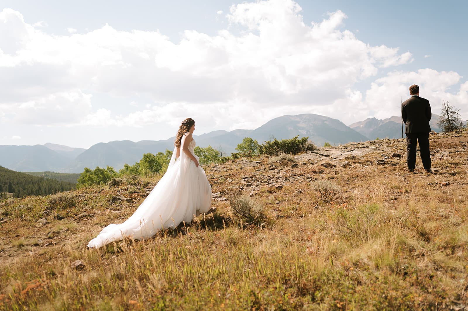 A woman in a white wedding dress walks along a dirt path through a grassy field under a clear sky.