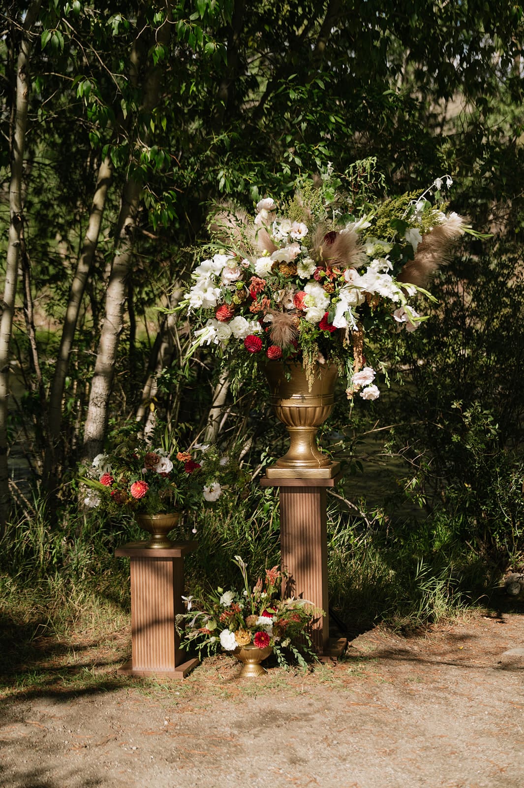 Tall gold vase with white and red flowers on a pedestal outdoors, surrounded by greenery and smaller floral arrangements at the base.