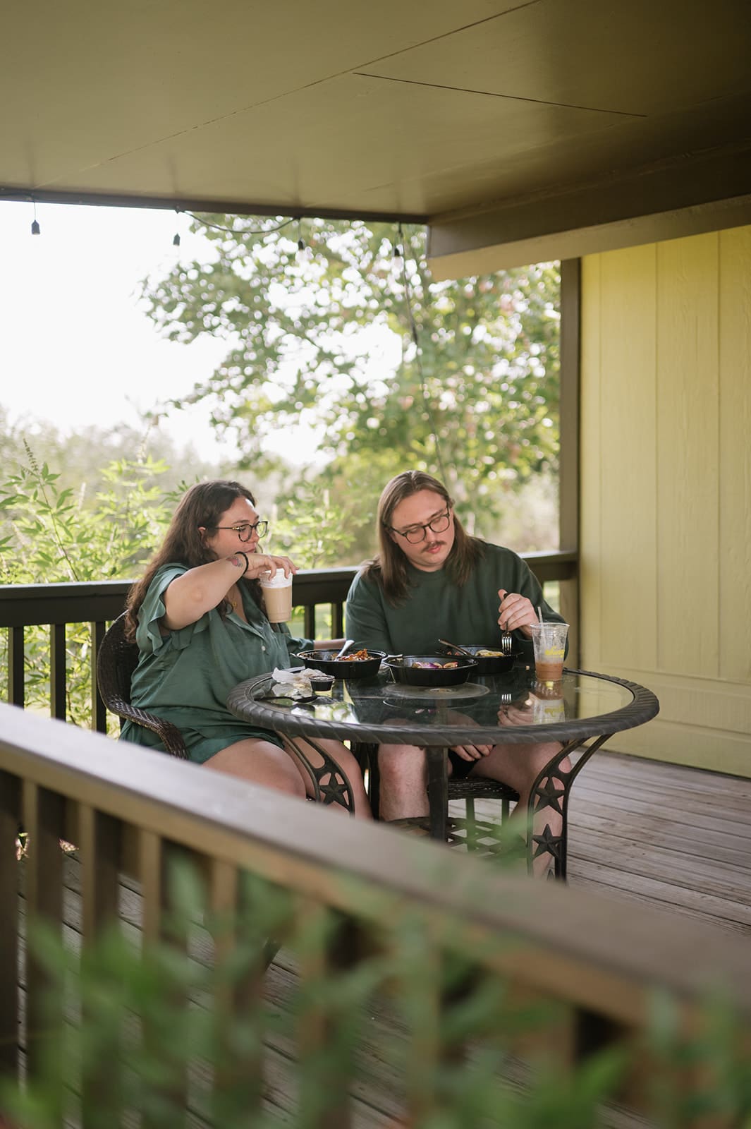 Two people sit at a round glass table on a wooden porch, eating and drinking together on a sunny day.