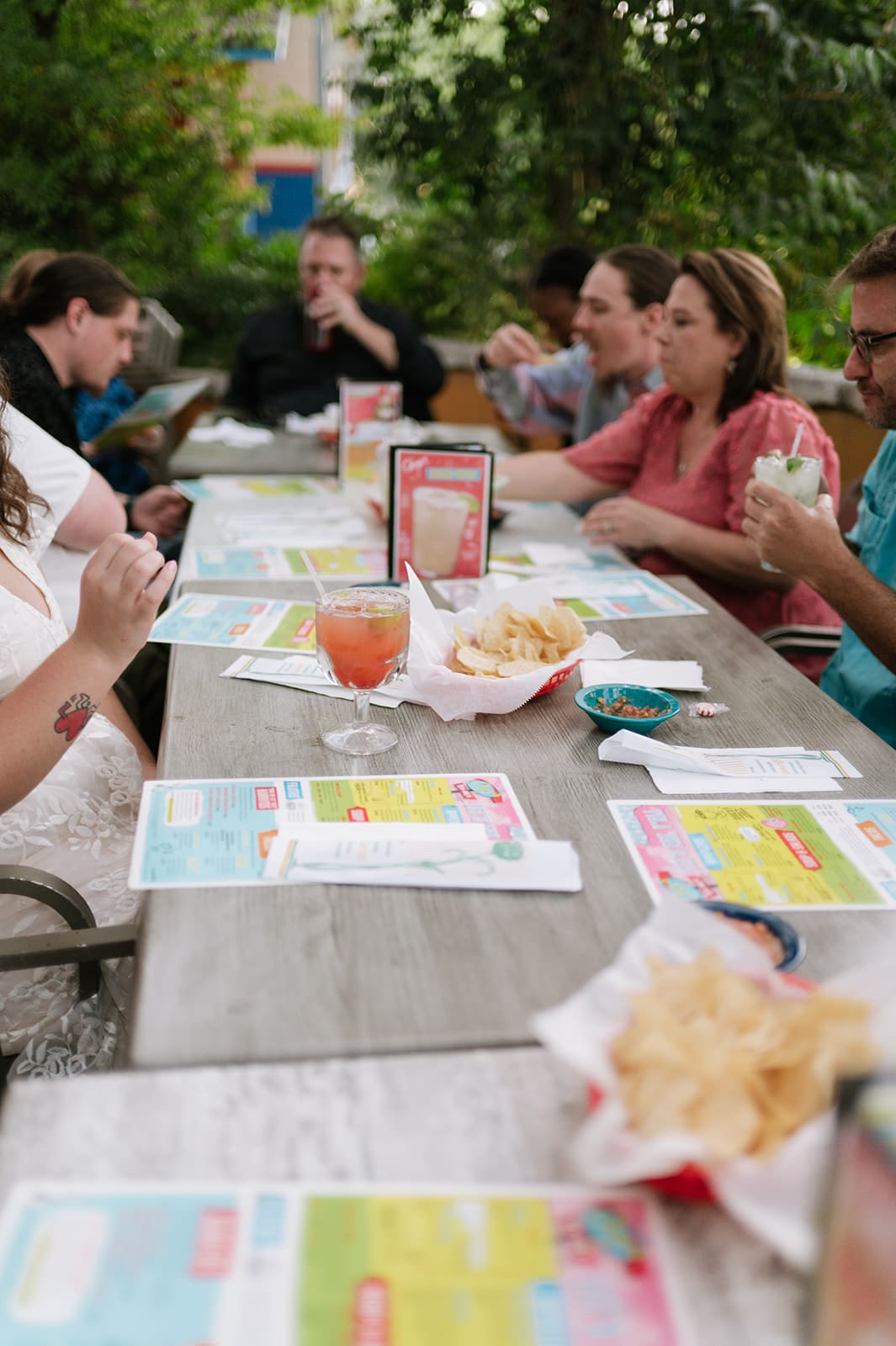 A group of twelve people sit around a long outdoor table at a restaurant, smiling for the camera, with menus and red cups in front of them.