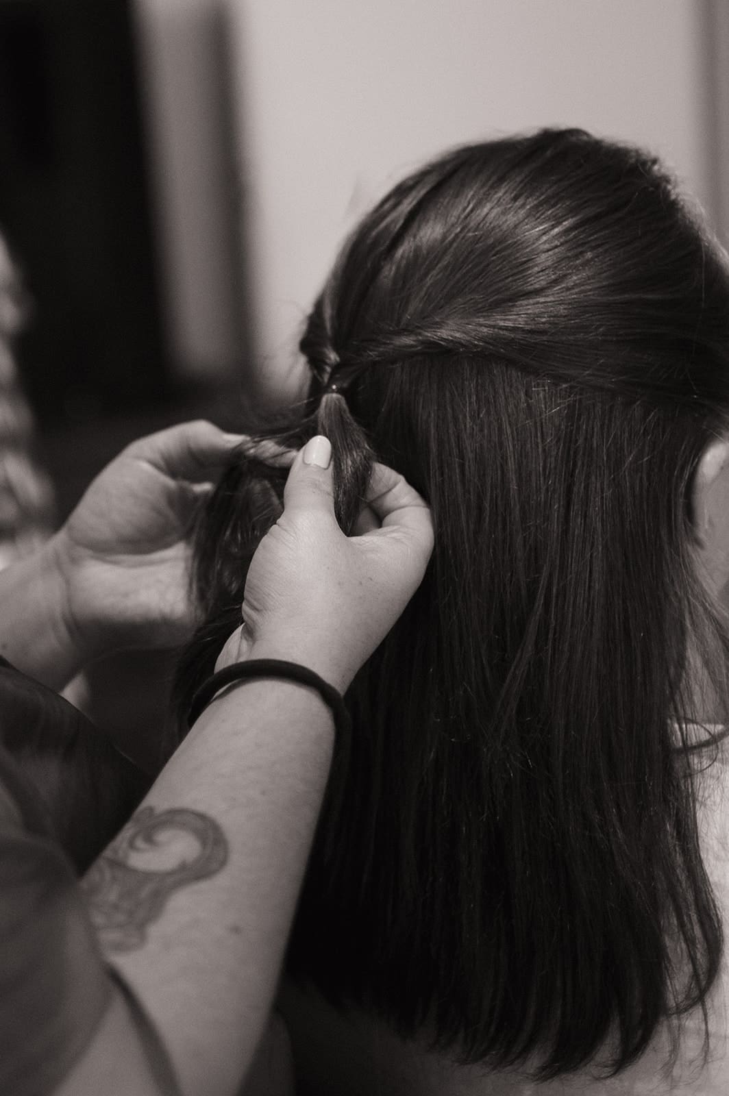 A woman sitting on a bed sprays a man's long hair while he sits in front of her with gold eye patches under his eyes.