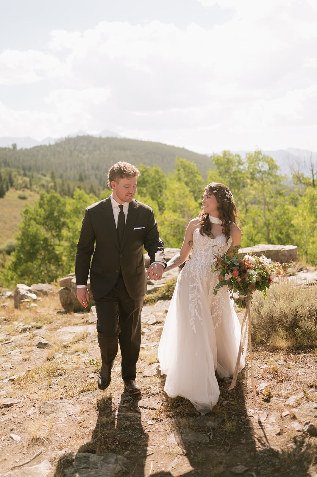 A bride and groom in wedding attire stand embracing on a rocky landscape with mountains and trees in the background under a partly cloudy sky for a Colorado wedding