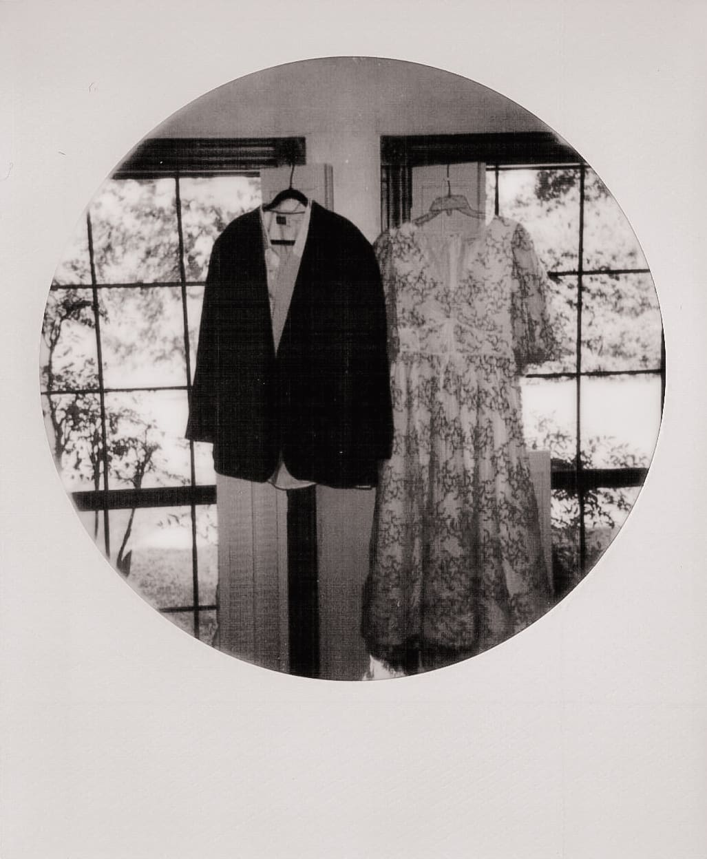 A suit and a floral dress are hanging side by side on hangers in front of large windows, with trees visible outside.