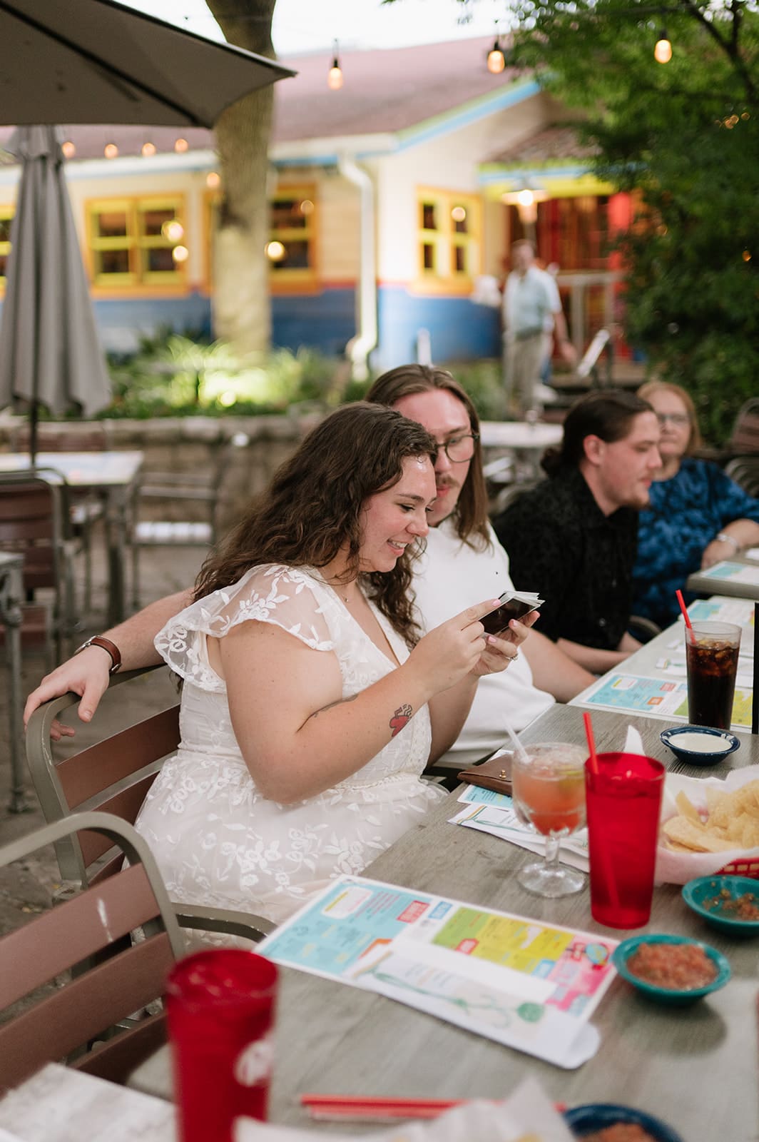 A group of twelve people sit around a long outdoor table at a restaurant, smiling for the camera, with menus and red cups in front of them.