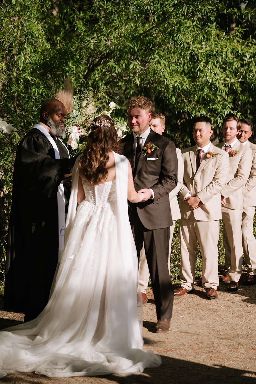A bride and groom stand together during an outdoor Colorado wedding ceremony as the officiant speaks; groomsmen in beige suits stand in the background 