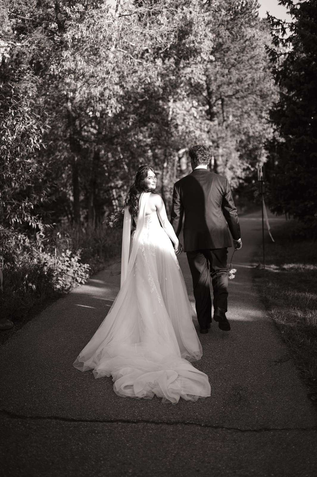 A bride and groom in wedding attire stand embracing on a rocky landscape with mountains and trees in the background under a partly cloudy sky for a Colorado wedding