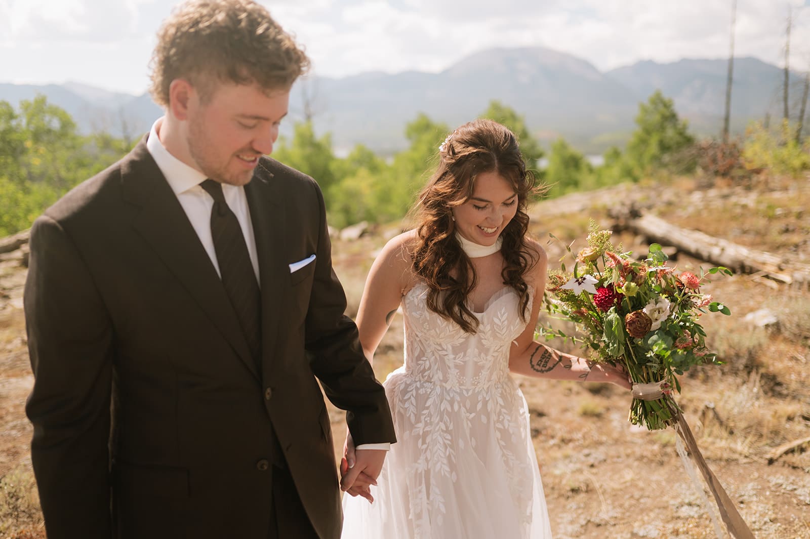 A bride and groom in wedding attire stand embracing on a rocky landscape with mountains and trees in the background under a partly cloudy sky for a Colorado wedding