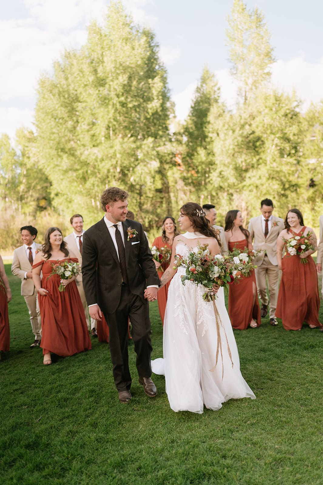 A bride and groom walk hand-in-hand outdoors, surrounded by their wedding party dressed in earth-toned attire, with trees in the background.