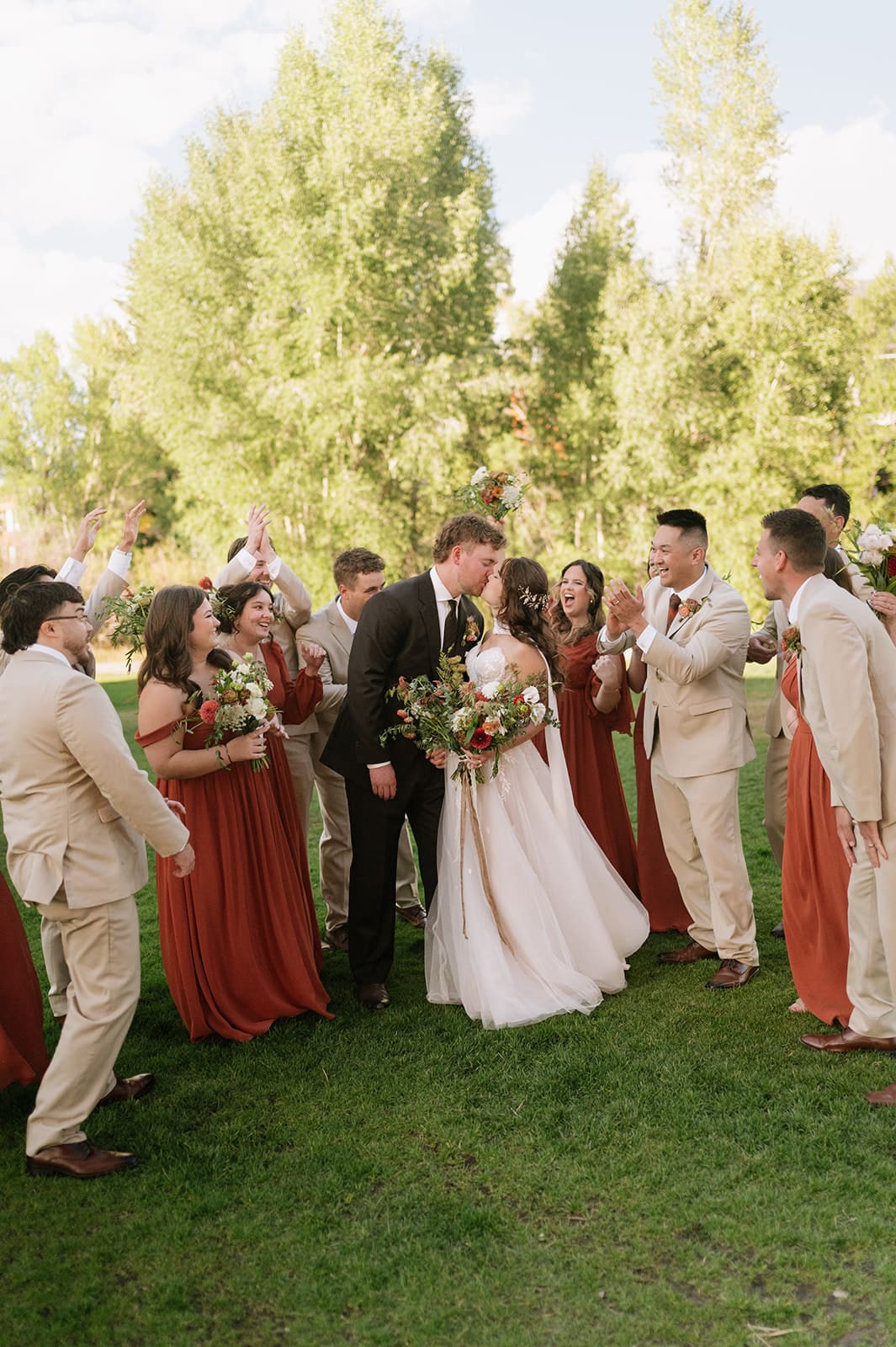 A bride and groom walk hand-in-hand outdoors, surrounded by their wedding party dressed in earth-toned attire, with trees in the background.
