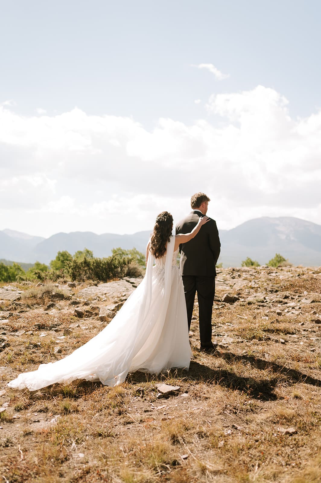 A bride and groom in wedding attire stand embracing on a rocky landscape with mountains and trees in the background under a partly cloudy sky for a Colorado wedding