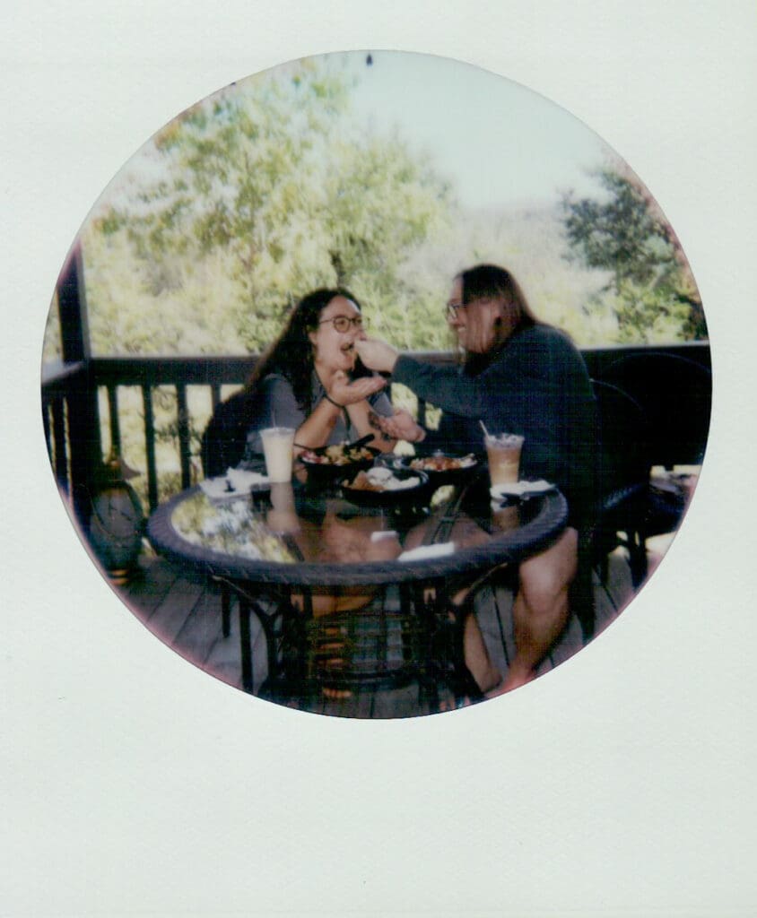 Two people sit at a round glass table on a wooden porch, eating and drinking together on a sunny day.