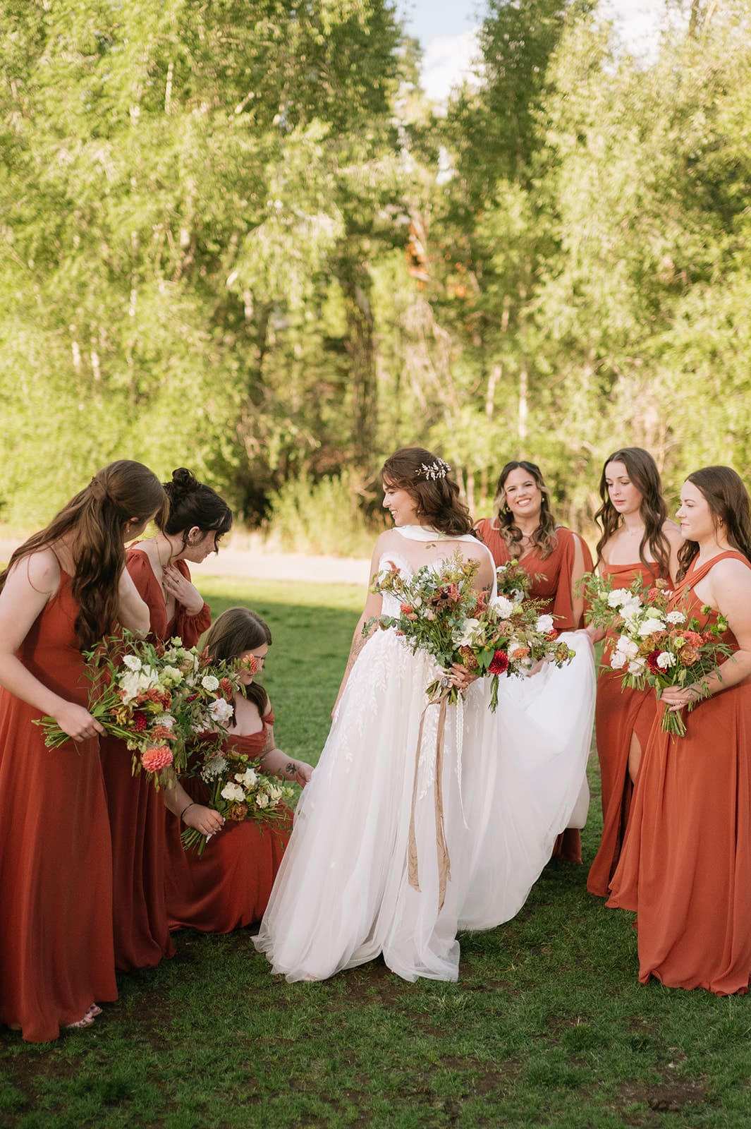 A bride and groom walk hand-in-hand outdoors, surrounded by their wedding party dressed in earth-toned attire, with trees in the background.