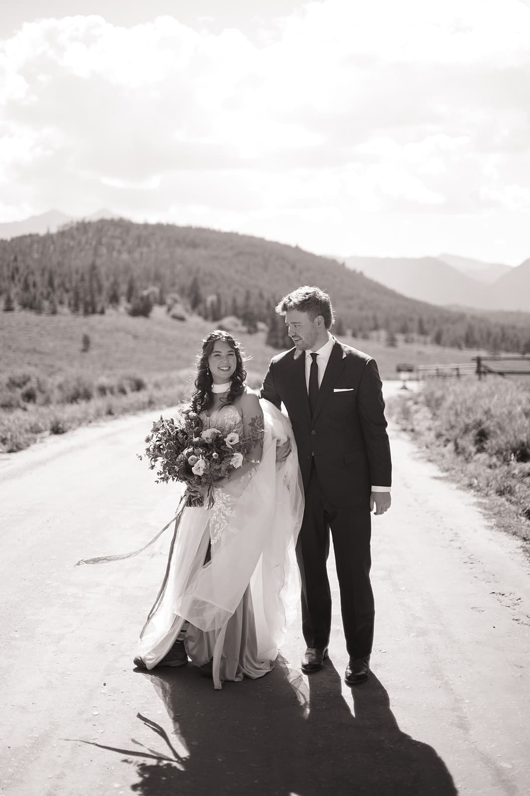 A bride and groom in wedding attire stand embracing on a rocky landscape with mountains and trees in the background under a partly cloudy sky for a Colorado wedding
