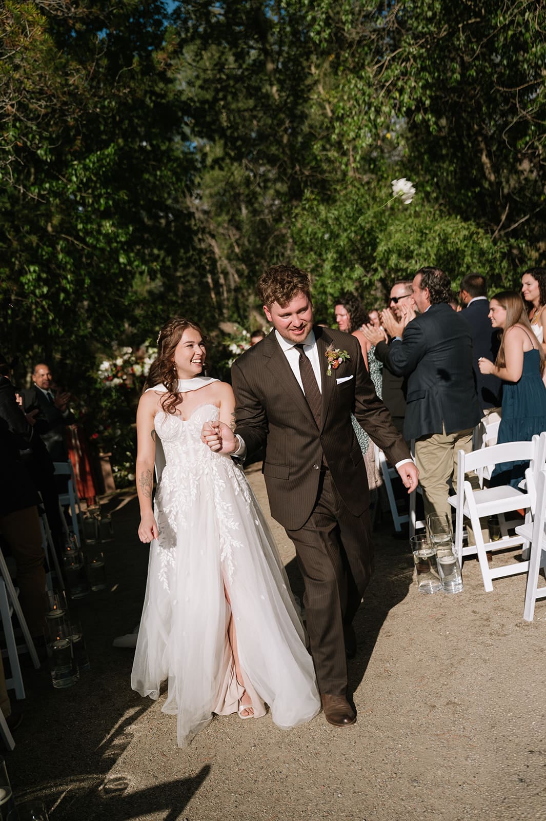 A bride and groom stand together during an outdoor Colorado wedding ceremony as the officiant speaks; groomsmen in beige suits stand in the background 