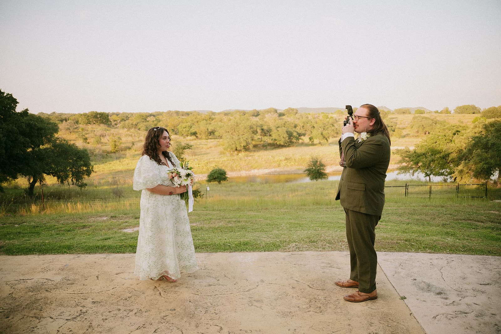A bride in a white dress holds flowers while a man in a suit takes her photo outdoors on a patio with a grassy landscape in the background for a texas hill country elopement