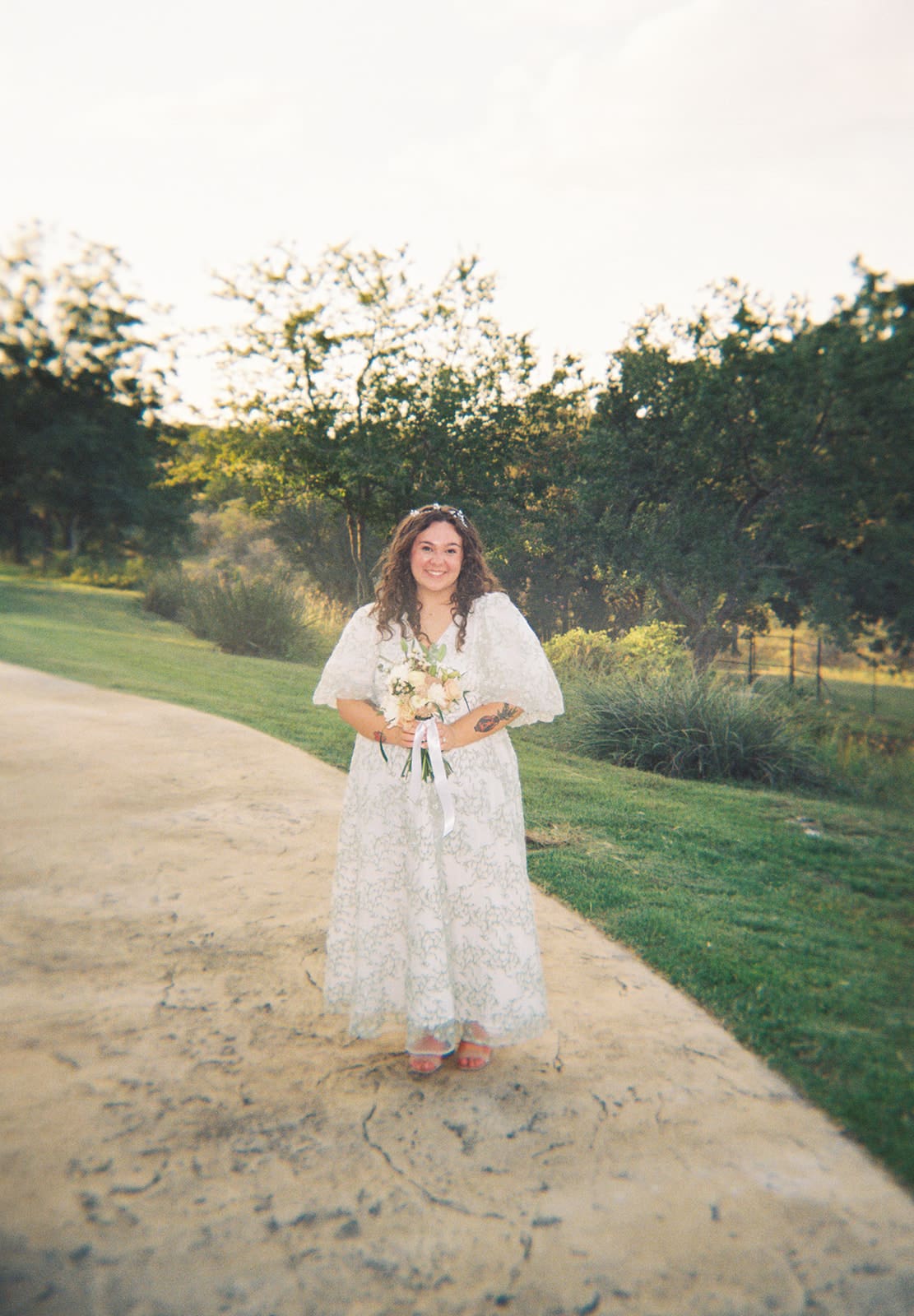 A woman in a white lace dress stands outdoors on a paved path, holding a bouquet, with trees and grass in the background.
