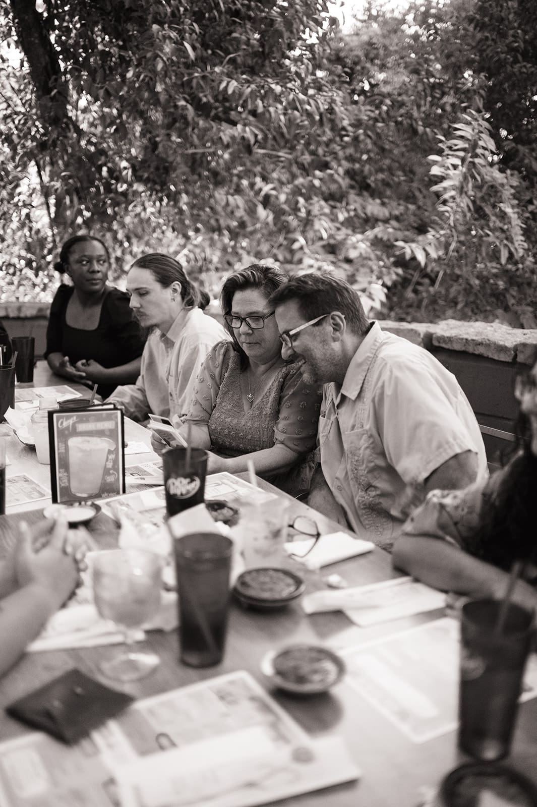 A group of twelve people sit around a long outdoor table at a restaurant, smiling for the camera, with menus and red cups in front of them.