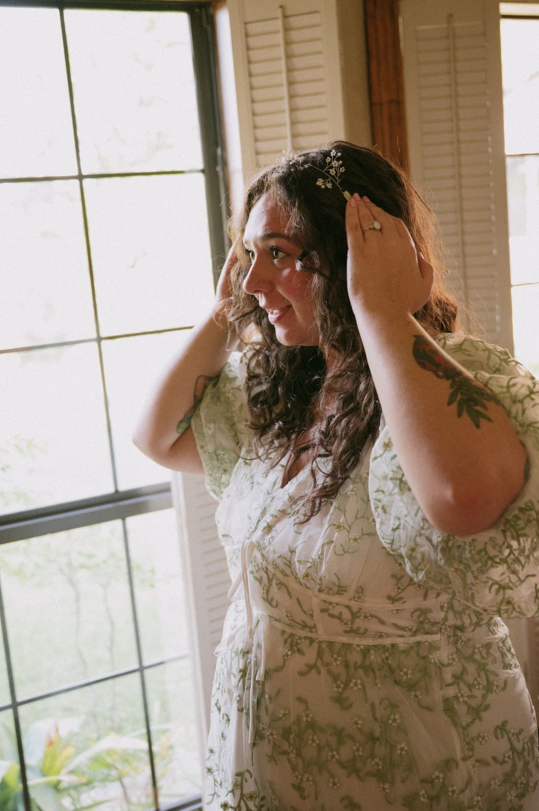 A woman in a light floral dress stands indoors by a window, adjusting her hair with both hands.