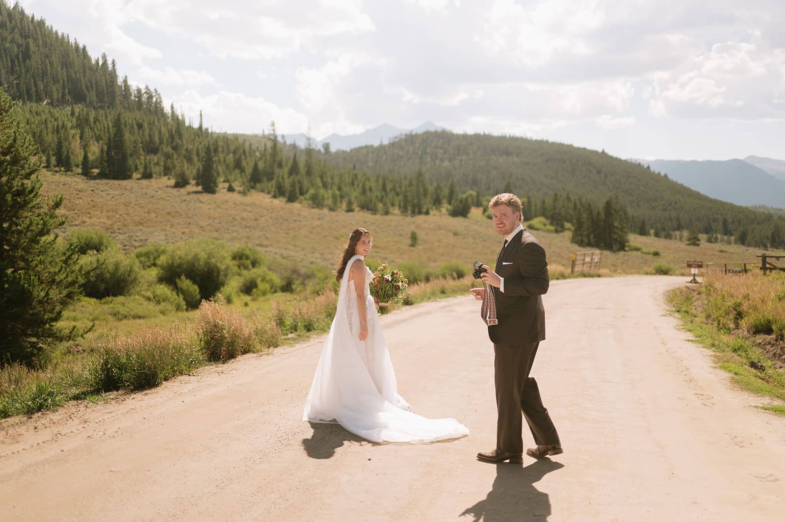 A bride and groom in wedding attire stand embracing on a rocky landscape with mountains and trees in the background under a partly cloudy sky for a Colorado wedding
