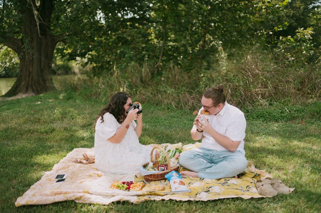 Two people sit on a blanket having a picnic outdoors, surrounded by greenery, with food and flowers arranged between them for a Texas Hill Country Elopement