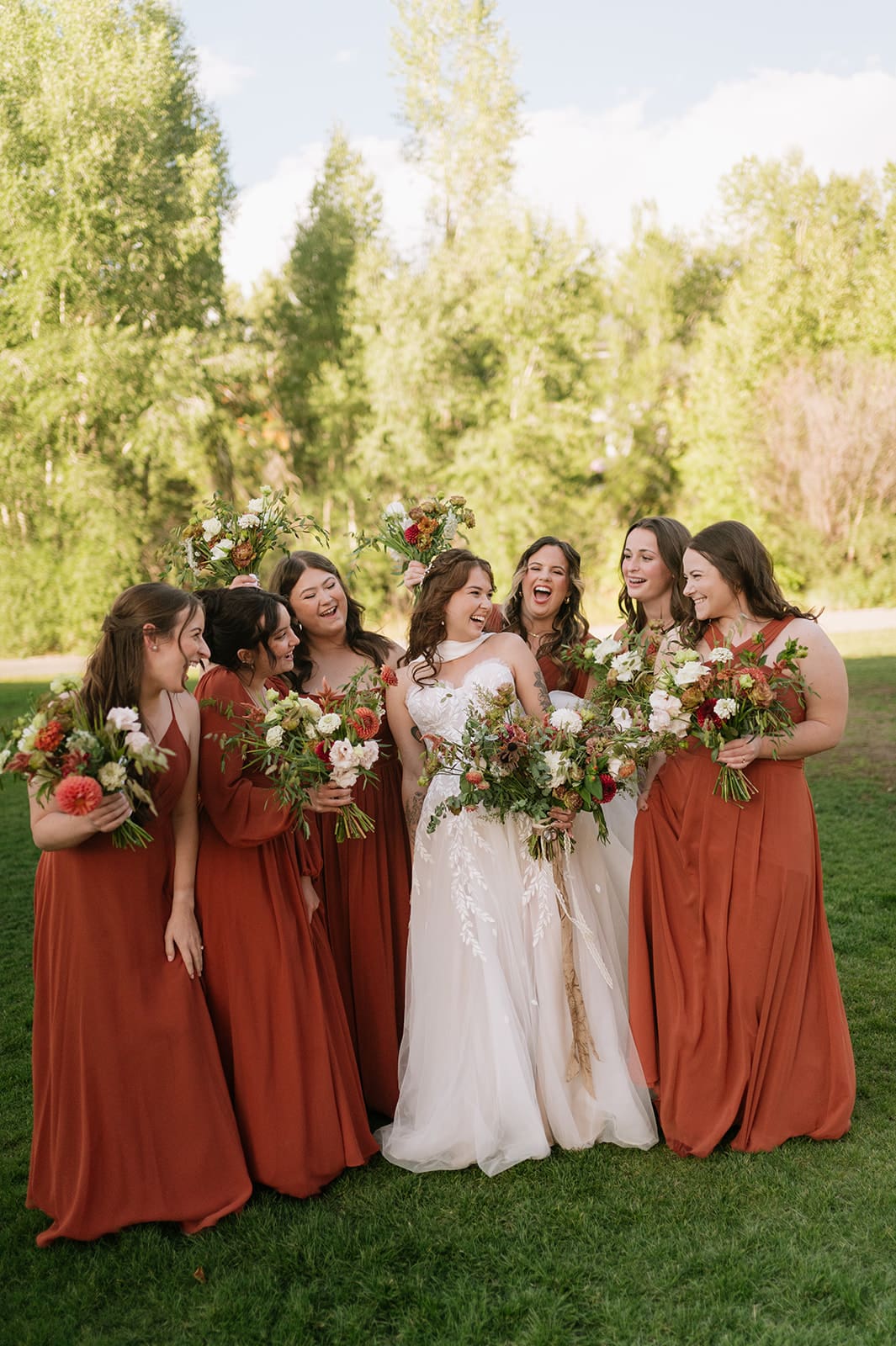 Five women stand together outdoors, one in a white wedding dress and four in matching rust-colored dresses, all holding bouquets of flowers and smiling.