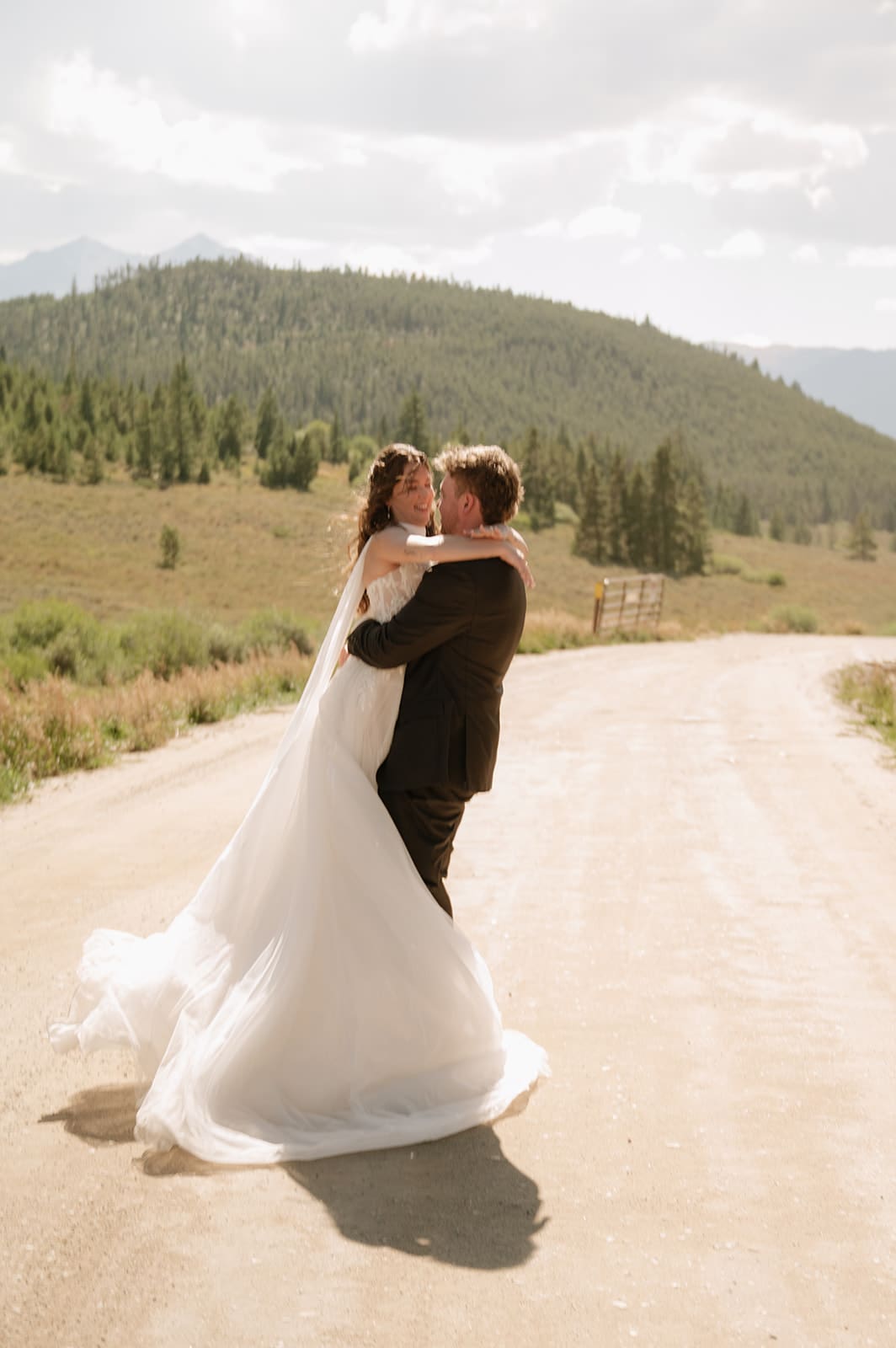 A bride and groom in wedding attire stand embracing on a rocky landscape with mountains and trees in the background under a partly cloudy sky for a Colorado wedding