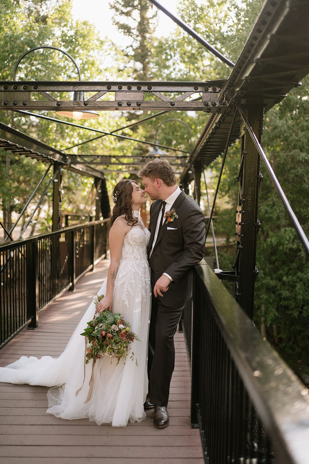 A bride in a white dress and a groom in a dark suit walk hand-in-hand on a metal bridge surrounded by trees.