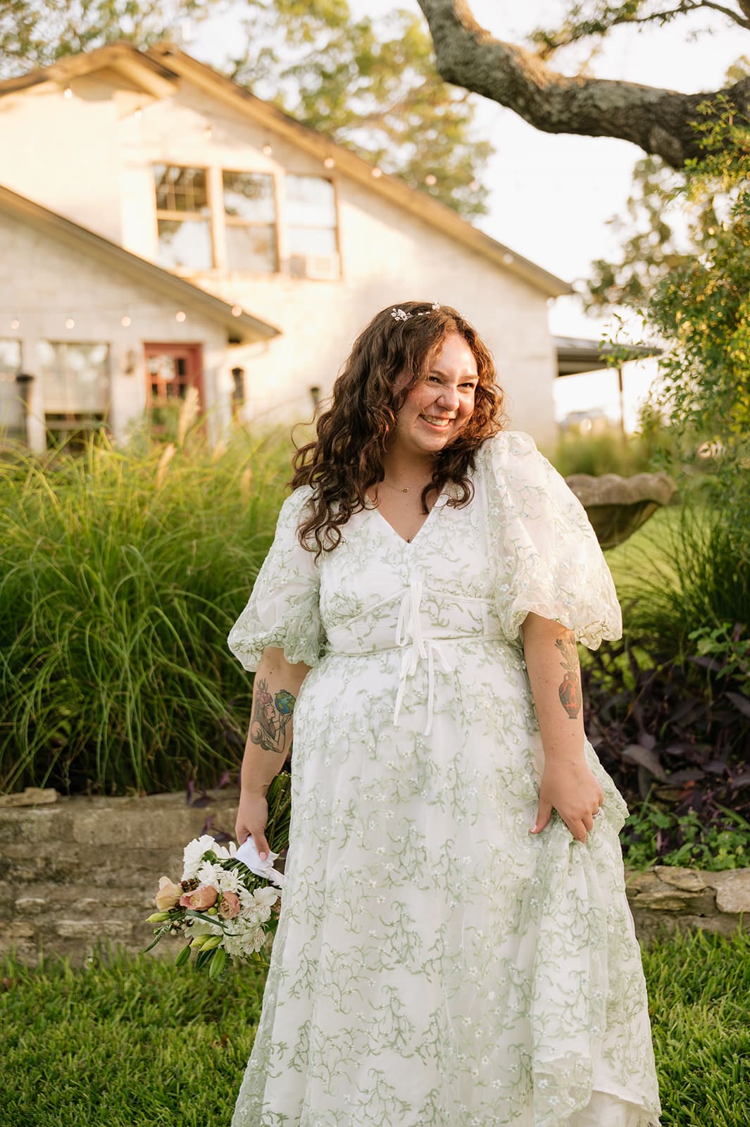 A woman in a white lace dress stands outdoors on a paved path, holding a bouquet, with trees and grass in the background.