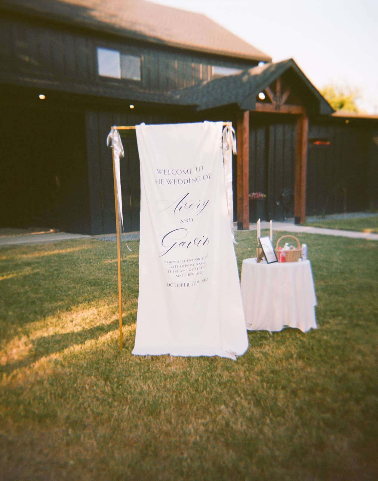 A white wedding welcome sign on a stand is displayed on grass near a small table with items, in front of a dark wooden building.