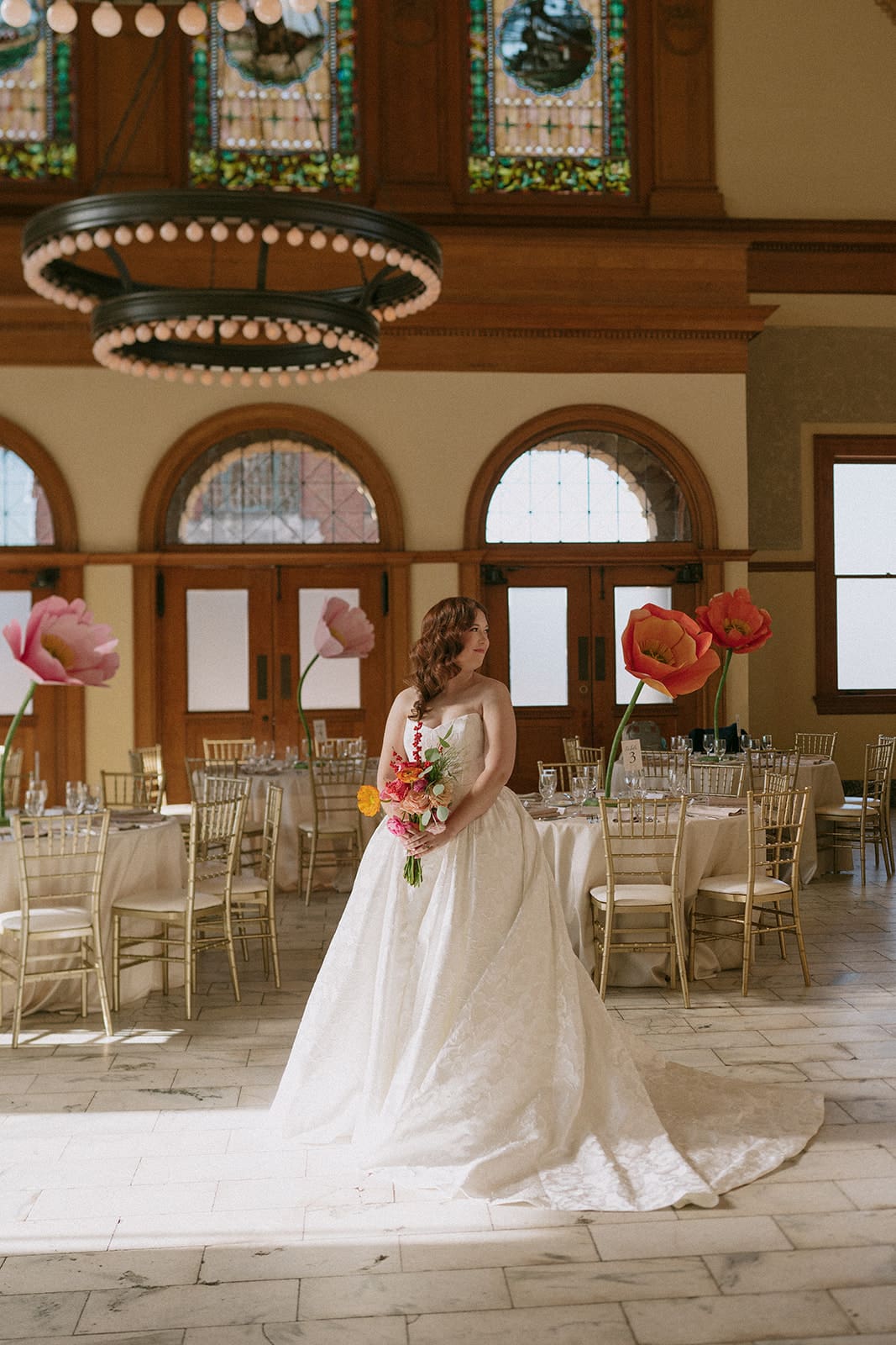 A bride in a white gown stands surrounded by seven bridesmaids in sage green dresses, all holding bouquets, inside a large, elegant hall with chandeliers at The Ashton Depot in Fort Worth