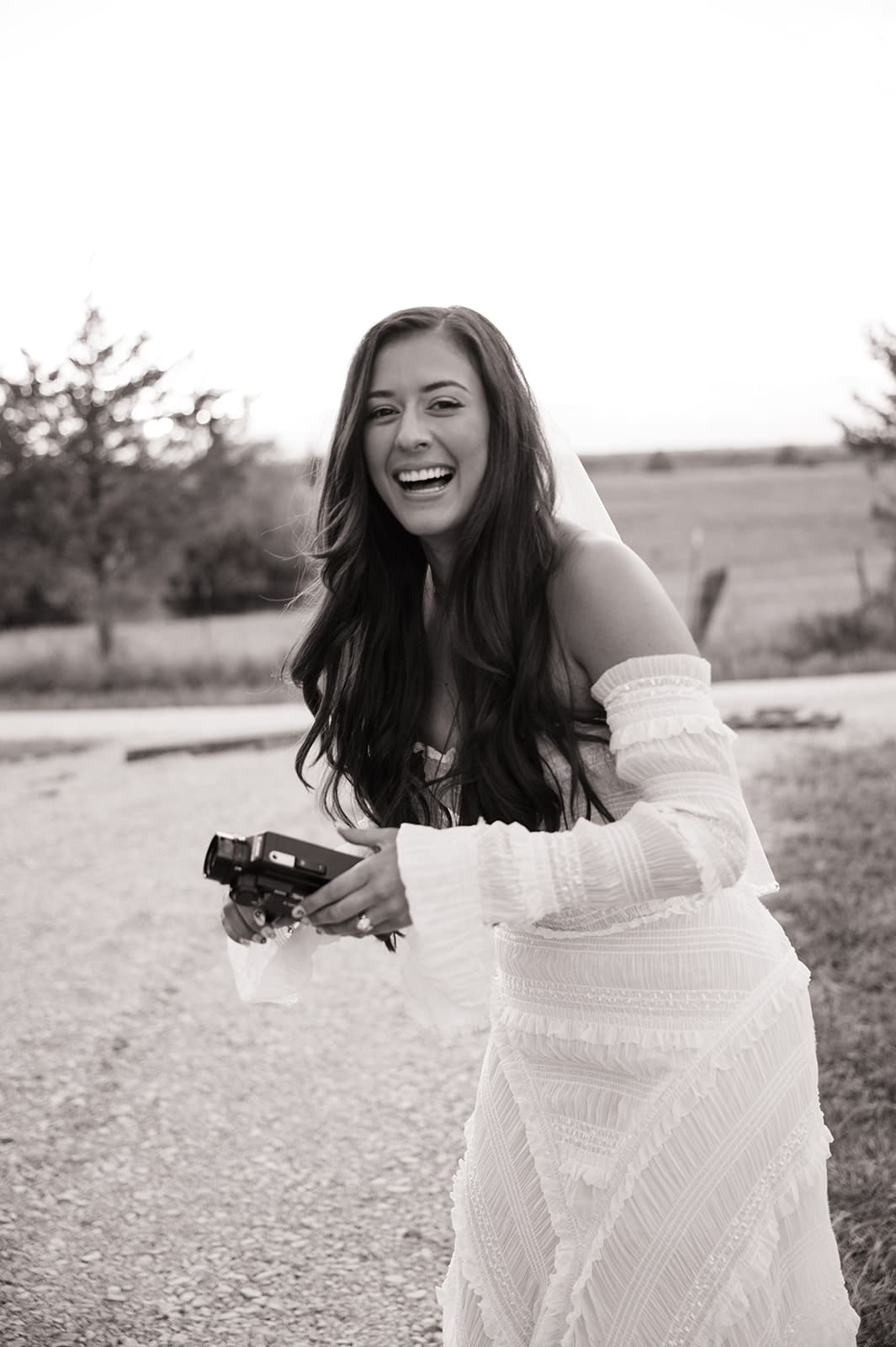 A woman in a wedding dress smiles and holds a camera outdoors on a gravel path, with fields and trees in the background.