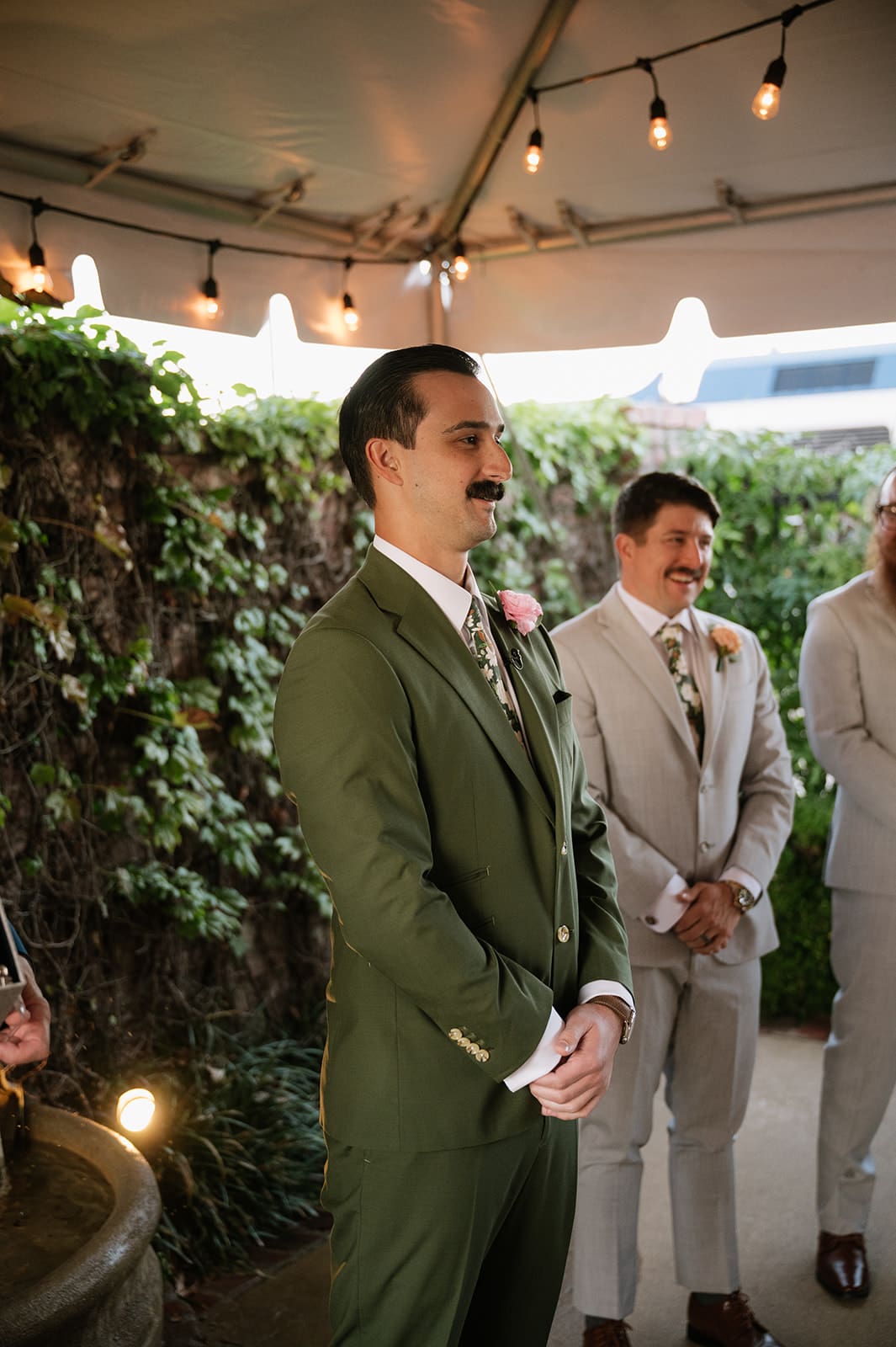 A man in a green suit stands under a tent, flanked by two men in light-colored suits, with string lights overhead and greenery in the background.