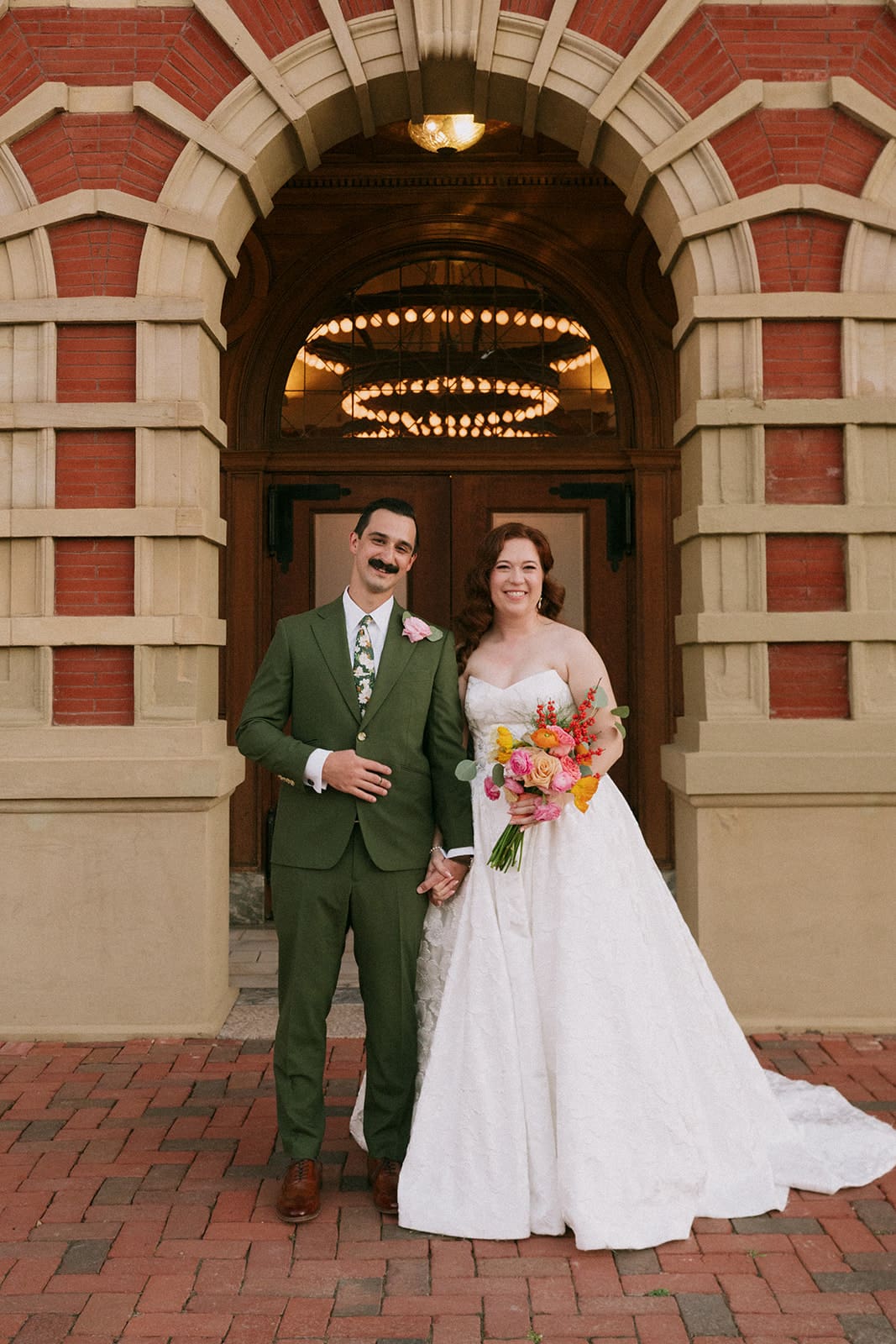 A bride in a white gown and a groom in a green suit stand holding hands and smiling in front of an arched brick building entrance for a wedding at The Ashton Depot in Fort Worth