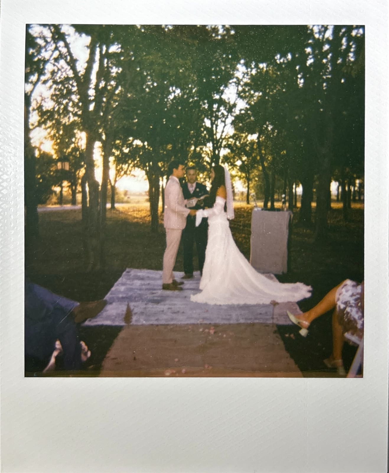 A bride and groom stand facing each other at an outdoor backyard wedding ceremony with trees and sunlight in the background