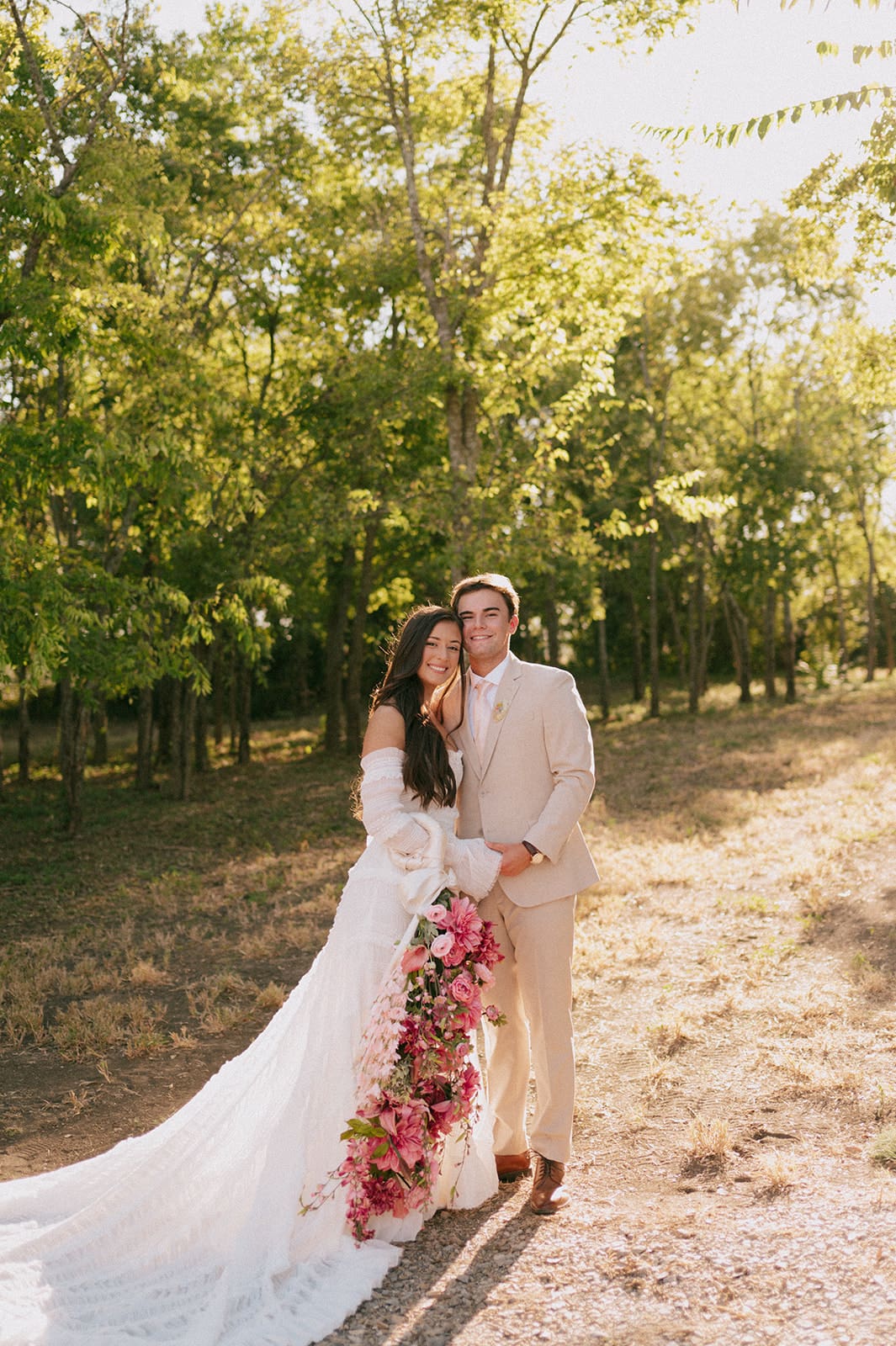 A bride and groom stand outdoors facing each other, holding hands in sunlight, with the bride holding a large bouquet of pink flowers for a backyard wedding in texas