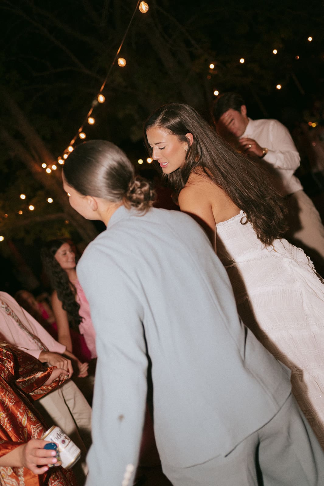 A group of people, including women in floral dresses, smile and dance outdoors at night under string lights