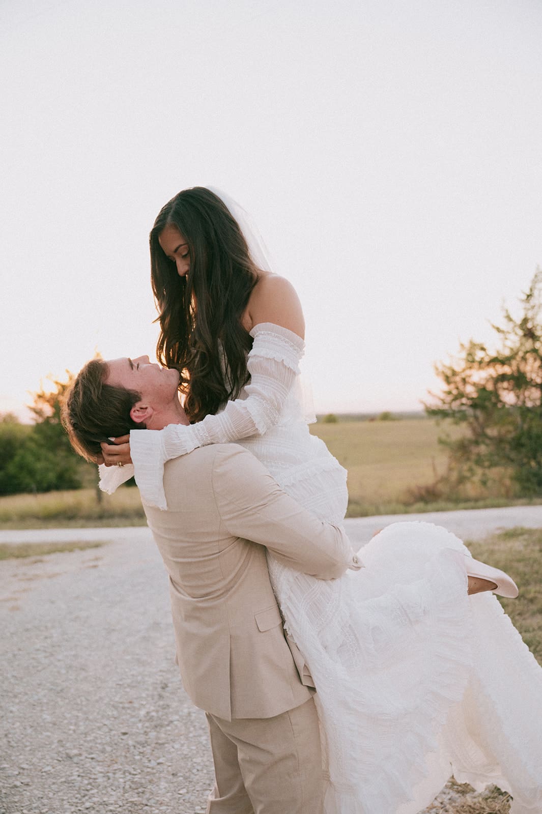 A groom in a beige suit lifts a bride in a white dress outdoors on a gravel path, with trees and fields in the background.