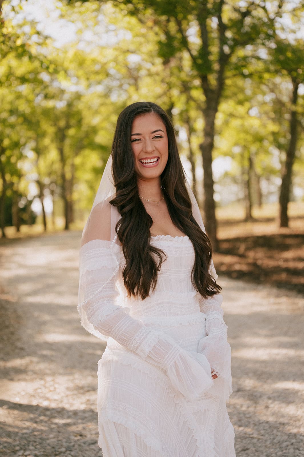 A bride in a wedding dress and veil hugs a man in a suit outdoors, surrounded by trees.