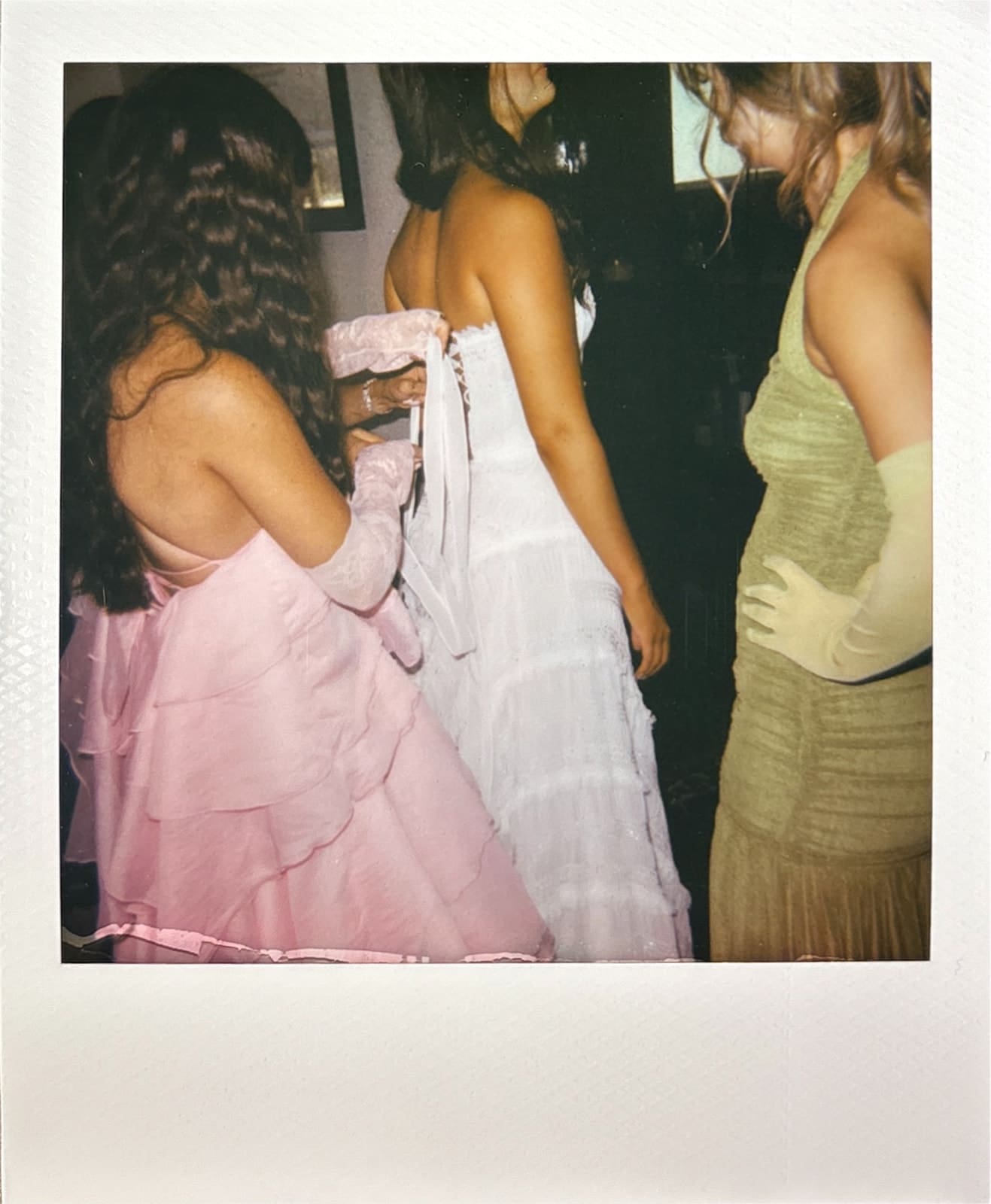 Three women in formal dresses stand together; one in pink helps another in white with the back of her dress, while the third, in a green dress and gloves, stands nearby.